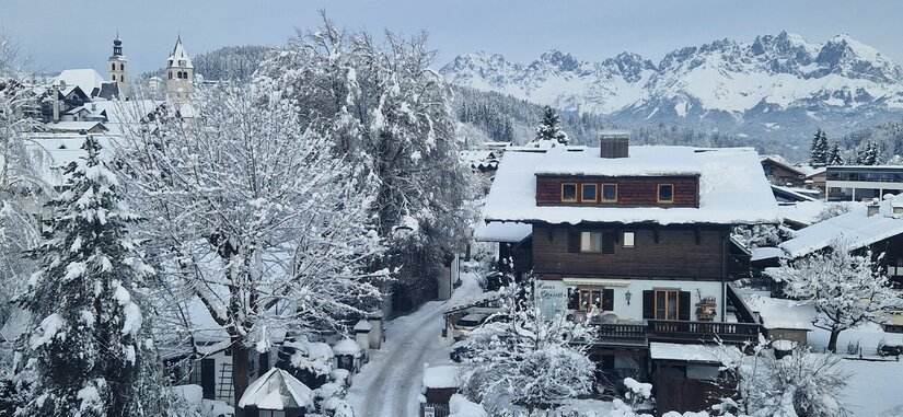 Verschneites Holzhaus in einem Alpendorf vor einer markanten, schneebedeckten Gebirgskette.