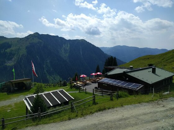 Hochwildalm mit Bergpanorama Berghütte mit Sonnenterrasse und Photovoltaikanlage vor bewaldeten Berghängen unter bewölktem Himmel.