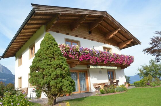 Haus Jöchl in Reith Zweistöckiges weißes Haus mit Holzdach, blumengeschmücktem Balkon und einer Terrasse mit Sitzgelegenheiten.