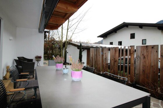 Terrasse mit großem Esstisch, Stühlen und Blumenvasen vor einem Wohnhaus.