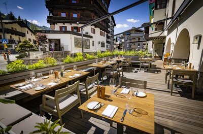Außenterrasse eines Restaurants mit eingedeckten Holztischen, Stühlen und Blick auf alpine Gebäude.