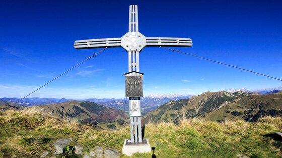 Ein metallisches Gipfelkreuz auf einem grasbewachsenen Bergkamm vor blauem Himmel.