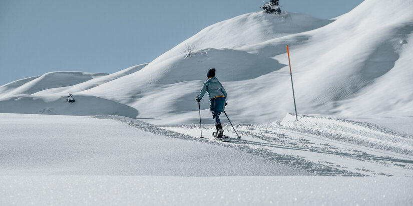 Eine Person wandert mit Schneeschuhen durch eine unberührte, schneebedeckte Landschaft unter klarem Himmel.