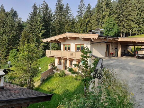 Zweistöckiges alpines Wohnhaus mit Holzbalkon vor einem dichten Nadelwald.