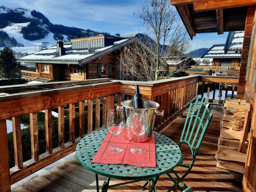 Holzbalkon mit rundem Tisch, zwei Weingläsern und Sektkühler vor alpiner Kulisse mit Chalets und Bergen.