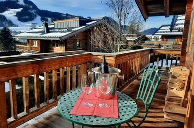 Holzbalkon mit rundem Tisch, zwei Weingläsern und Sektkühler vor alpiner Kulisse mit Chalets und Bergen.