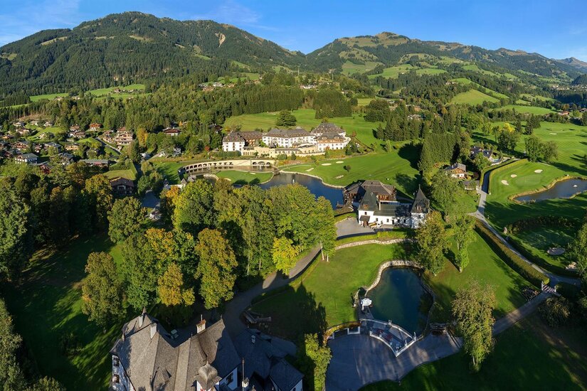 Luftaufnahme eines Golfplatzes mit Clubhaus, Teichen und bewaldeten Bergen im Hintergrund.