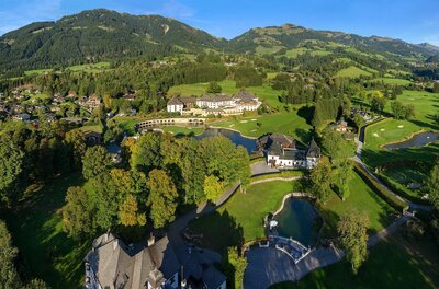 Luftaufnahme eines Golfplatzes mit Clubhaus, Teichen und bewaldeten Bergen im Hintergrund.