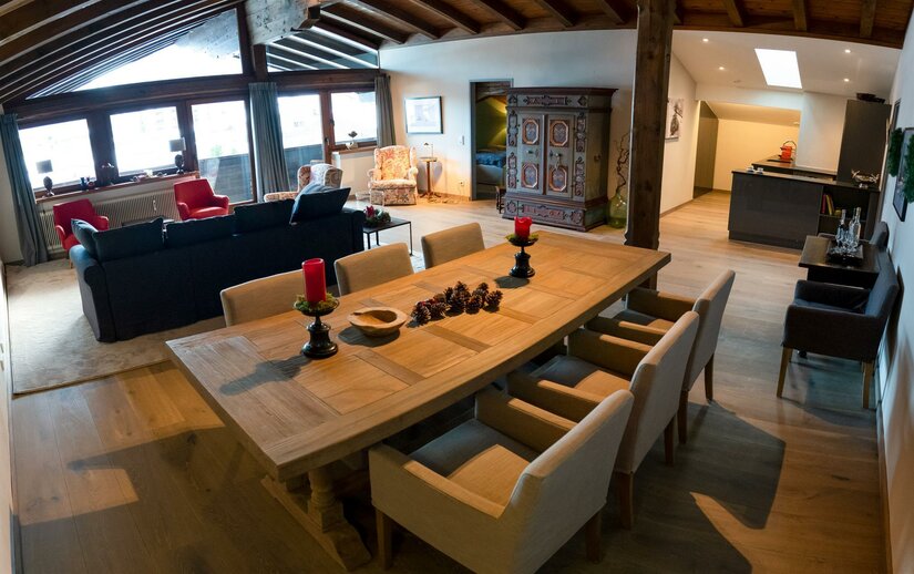 Moderner Wohn- und Essbereich mit großem Holztisch, dunklem Sofa und rustikalem Schrank unter einer Dachschräge.