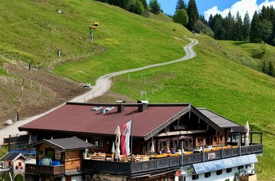 Berghütte mit Terrasse vor einem grünen Hang und einem geschwungenen Wanderweg.