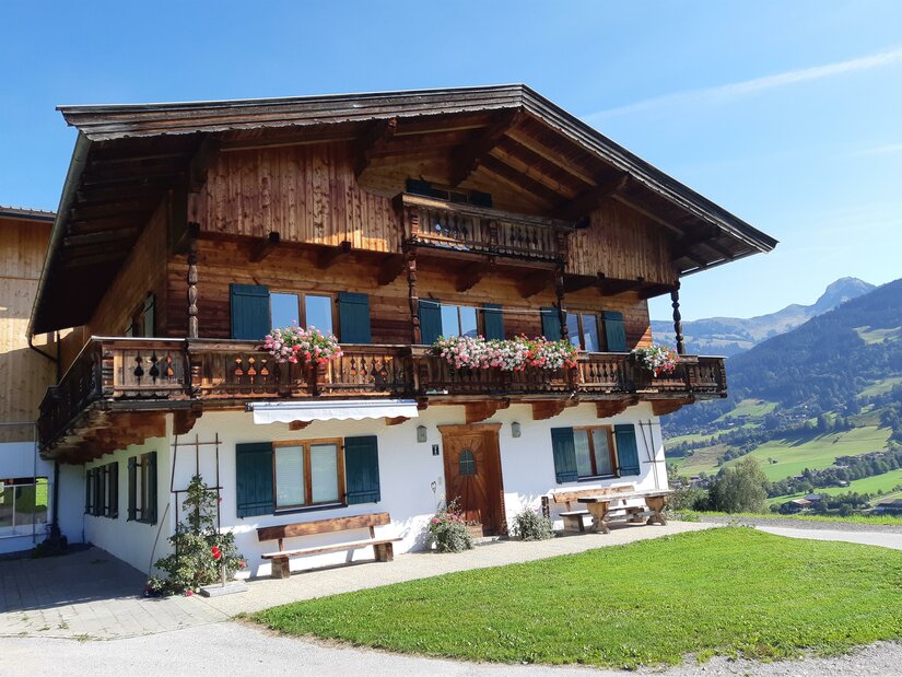 Traditionelles Alpenhaus aus Holz und Stein mit Blumenbalkonen vor Gebirgspanorama.