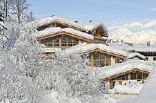 Verschneite Holzchalets mit schneebedeckten Dächern und Bäumen vor Gebirgskulisse.