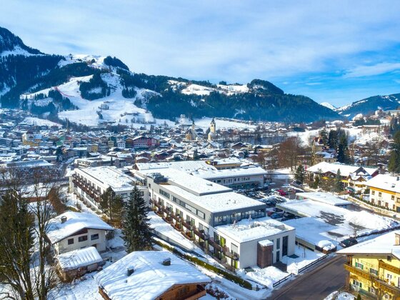 Luftaufnahme der Rehaklinik Kitzbühel und des verschneiten Ortes vor bewaldeten Bergen.
