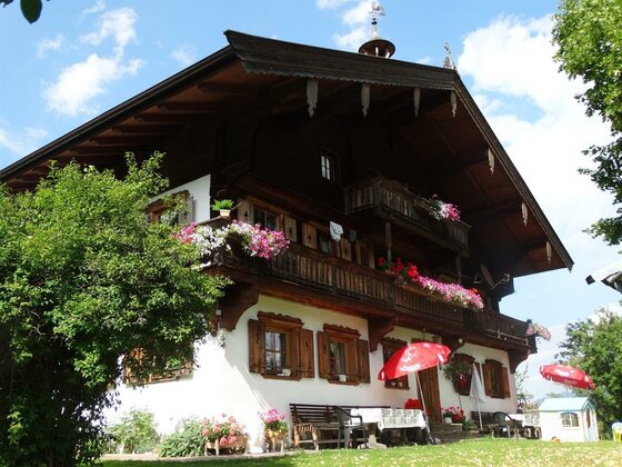 Hausansicht Plattenhof Alpenländisches Bauernhaus mit Holzbalkonen, Blumenschmuck und Außenterrasse unter blauem Himmel.