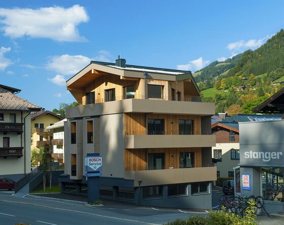 Modernes mehrstöckiges Gebäude mit Holzfassade und Balkonen vor bewaldeten Bergen.