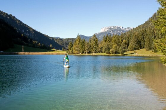 Ein Mann paddelt auf einem Stand-Up-Paddleboard auf einem ruhigen Gewässer. Im Hintergrund sind grüne Berge und ein klarer blauer Himmel zu sehen.