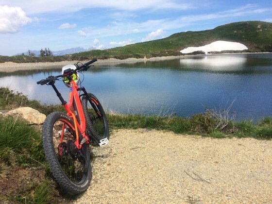 Mountainbike am Hahnenkamm-See Ein rotes Mountainbike steht auf einem Schotterweg vor einem Bergsee mit Schneeresten am Ufer.