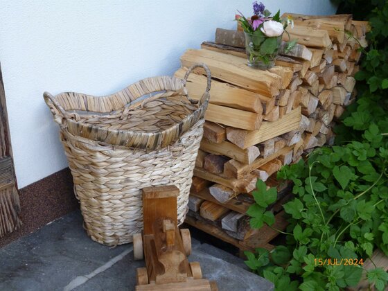 Ein geflochtener Korb, ein Holzspielzeug und aufgestapeltes Brennholz mit Blumenvase vor einer weißen Wand.