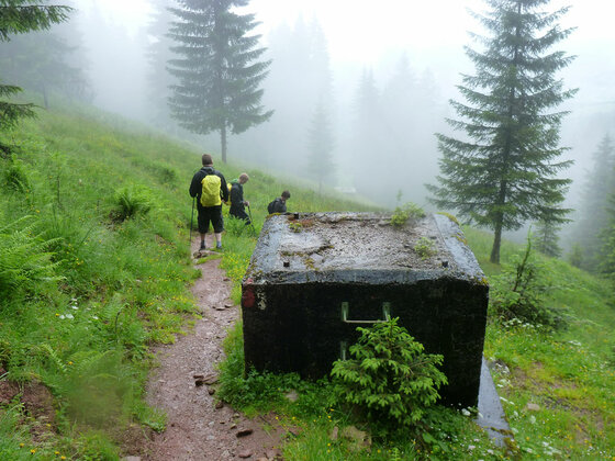 Wanderer auf einem schmalen Pfad im Nebel neben einem Betonbauwerk.
