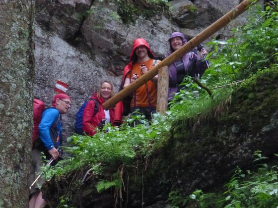 Vier Wanderer mit Regenbekleidung und Wanderstöcken auf einem schmalen Pfad an einer Felswand.