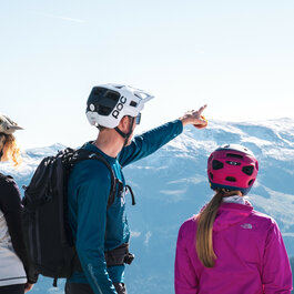 Drei Radfahrer stehen in Kitzbühel auf einem Berg und blicken auf die schneebedeckten Gipfel in der Ferne, während einer auf einen Punkt zeigt.