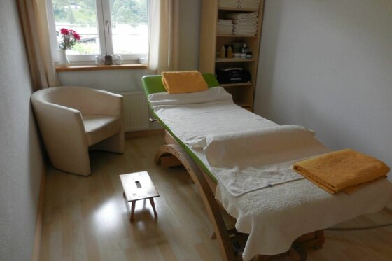 Massageliege mit weißen und gelben Handtüchern in einem hellen Behandlungsraum mit Sessel und Fenster.