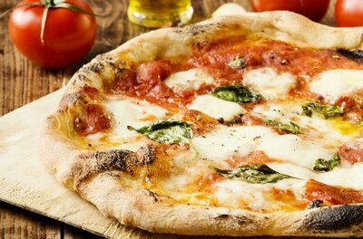Eine frisch gebackene Pizza mit Mozzarella, Tomatensauce und frischem Basilikum. Im Hintergrund sind frische Tomaten und Olivenöl zu sehen.