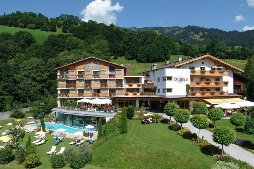 Außenansicht des Hotels Bichlhof mit Außenpool, Liegewiese und bewaldeten Bergen im Hintergrund.