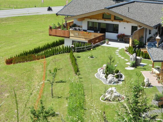 Ferienwohnung Widmann Jochberg Einfamilienhaus mit gepflegtem Garten, Steingarten-Elementen und Balkon neben einer Landstraße.