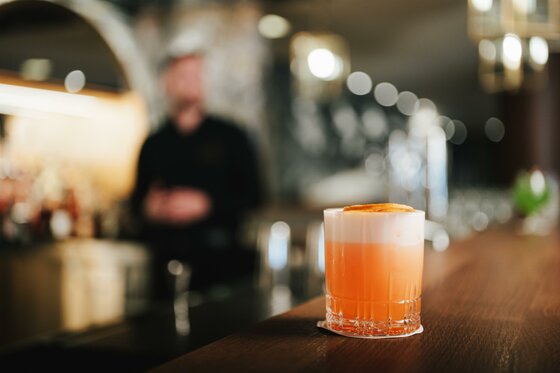 Cocktail in der Cooper Bar Ein orangefarbener Cocktail mit Schaumkrone auf einem Holztresen vor unscharfem Bar-Hintergrund.