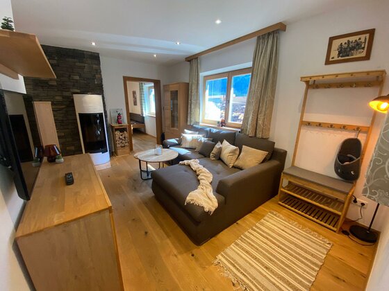 Ein gemütliches Wohnzimmer mit einem grauen Sofa, einem Tisch und einem modernen Fernseher. Große Fenster sorgen für viel Tageslicht und eine warme Atmosphäre.