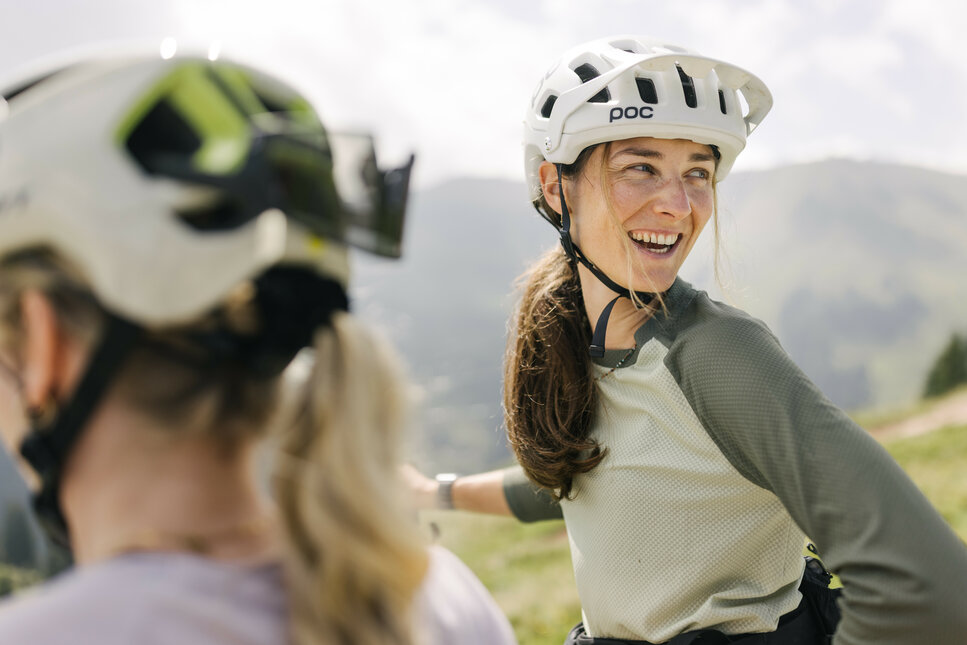 In Kitzbühel genießt eine Radfahrerin mit Helm die Berglandschaft und unterhält sich mit Freunden.