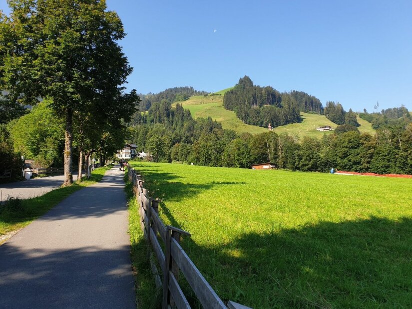 Asphaltierter Wanderweg neben einer grünen Wiese mit Holzzaun und bewaldeten Hügeln im Hintergrund.