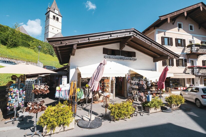Kitzbüheler Ladl Souvenir Souvenirgeschäft mit Außenverkaufsständern vor einem weißen Gebäude mit Holzdach und Kirchturm im Hintergrund.