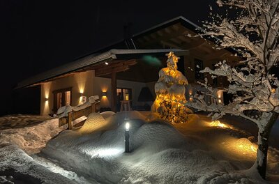 Beleuchtetes Holzhaus bei Nacht mit tief verschneitem Garten und einem geschmückten Tannenbaum.