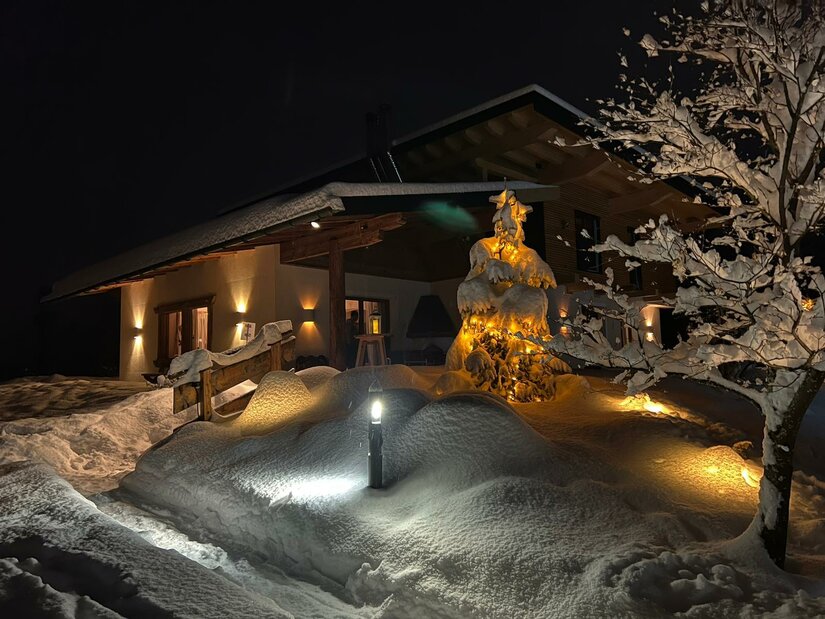 Haus Skida im Winter Beleuchtetes Holzhaus bei Nacht mit tief verschneitem Garten und einem geschmückten Tannenbaum.