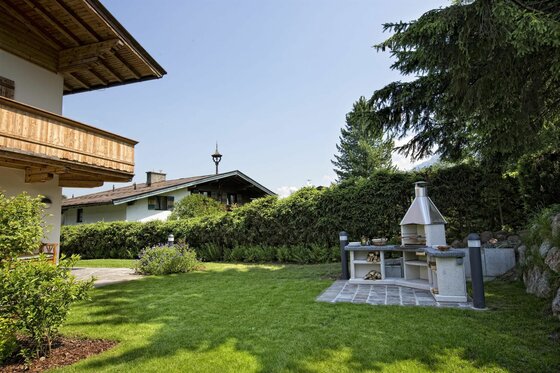 Gartenanlage mit gemauertem Außenkamin, Rasenfläche und angrenzendem Landhaus unter blauem Himmel.