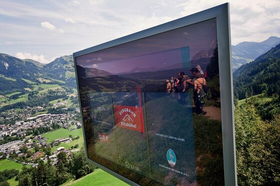 Ein Monitor mit Informationen der Bergbahn Kitzbühel vor einer alpinen Bergkulisse mit Blick auf ein Tal.