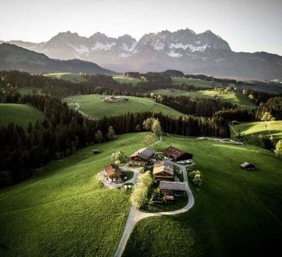Hof Hintererb Kitzbühel Luftansicht Luftaufnahme eines Bauernhofs auf einer grünen Wiese vor einem bewaldeten Tal und schroffen Berggipfeln.