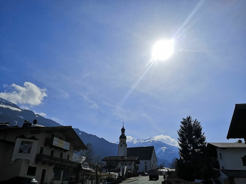 Ortsansicht Hechenmoos mit Kirche Blick auf eine Dorfkirche mit Zwiebelturm vor schneebedeckten Bergen unter strahlender Sonne.