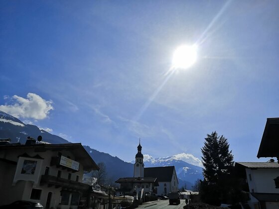 Ortsansicht Hechenmoos mit Kirche Blick auf eine Dorfkirche mit Zwiebelturm vor schneebedeckten Bergen unter strahlender Sonne.