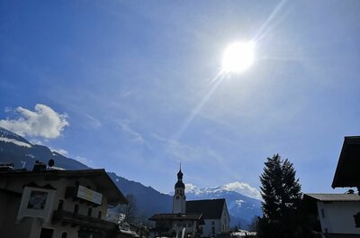 Ortsansicht Hechenmoos mit Kirche Blick auf eine Dorfkirche mit Zwiebelturm vor schneebedeckten Bergen unter strahlender Sonne.