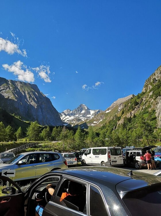 Parkplatz am Großglockner Parkplatz mit Autos vor bewaldeten Hängen und schneebedeckten Berggipfeln unter blauem Himmel.
