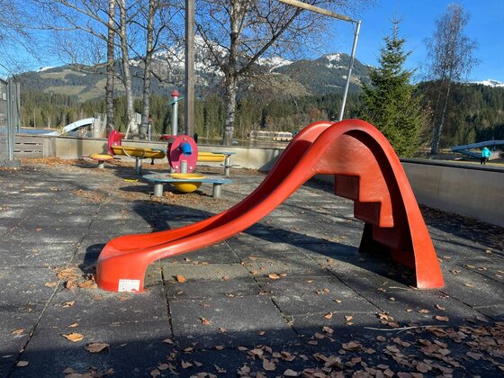 Spielplatz beim Schwarzsee Stadtbad Rote Kinderrutsche auf einem Spielplatz mit Gummiboden vor bewaldeten Bergen.