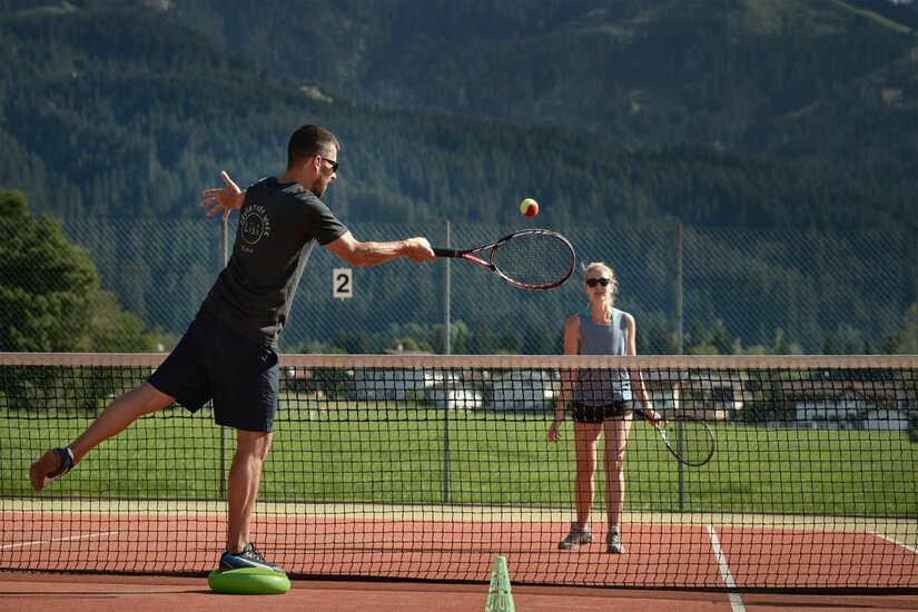 Tennis im Lisi Family Hotel Zwei Personen beim Tennisspielen auf einem Außenplatz vor bewaldeten Bergen.