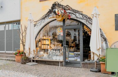 Eingangsbereich eines Geschäfts mit Rundbogenfenster, Keramikwaren im Schaufenster und zwei weißen Sonnenschirmen.