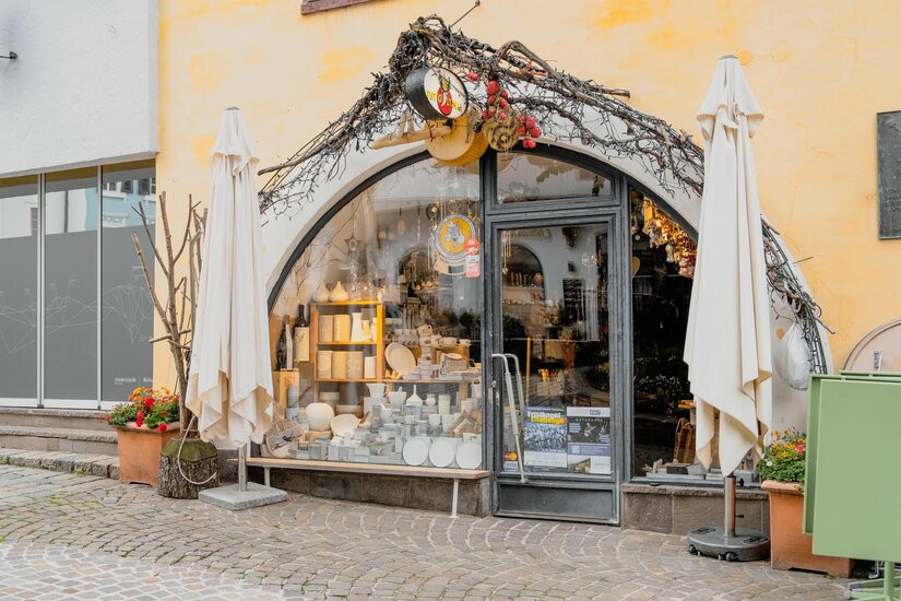 Eingangsbereich eines Geschäfts mit Rundbogenfenster, Keramikwaren im Schaufenster und zwei weißen Sonnenschirmen.