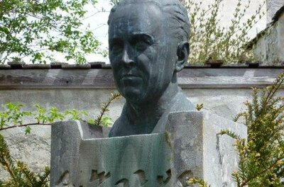 Eine Büste eines Mannes steht auf einem Steinsockel. Um die Skulptur herum sind grüne Pflanzen und Bäume zu sehen.