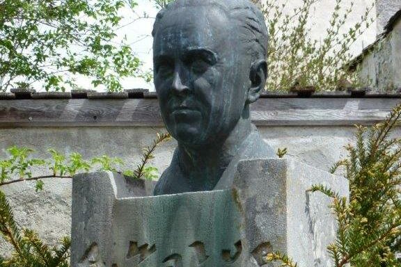 Eine Büste eines Mannes steht auf einem Steinsockel. Um die Skulptur herum sind grüne Pflanzen und Bäume zu sehen.