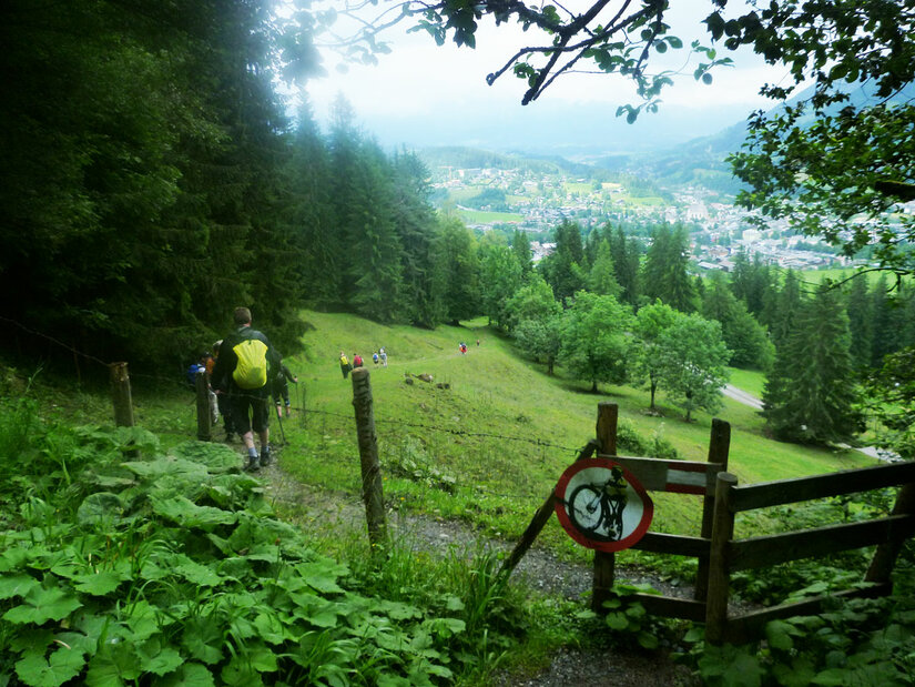 Wanderer auf einem schmalen Pfad neben einem Holzzaun mit Blick auf eine Wiese und ein Tal.